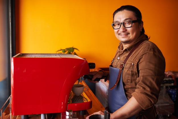 Free photo smiling middle-aged asian man in apron standing next to espresso machine and smiling