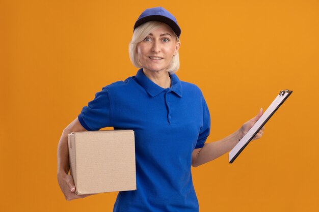 Улыбающаяся белокурая женщина-доставщик средних лет в синей униформе и кепке держит картонную коробку и буфер обмена