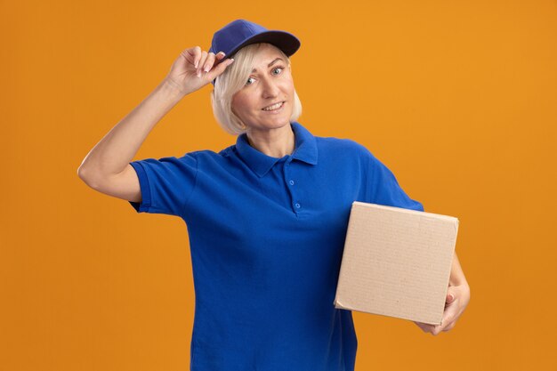 Улыбающаяся белокурая курьерская женщина средних лет в синей форме и кепке держит картонную коробку и хватает ее кепку
