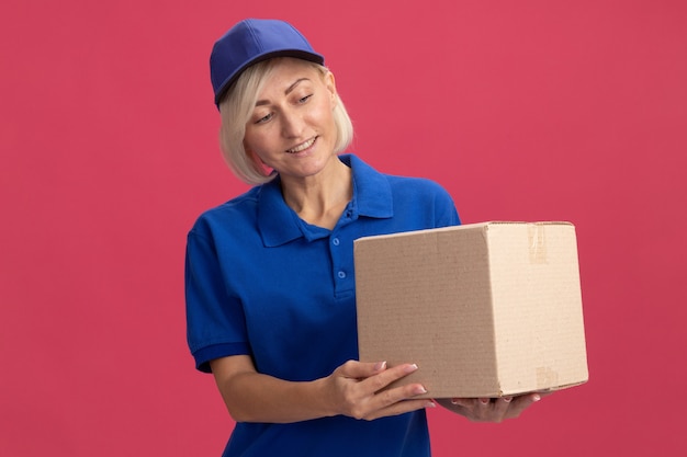Smiling middle-aged blonde delivery woman in blue uniform and cap holding and looking at cardboard box 