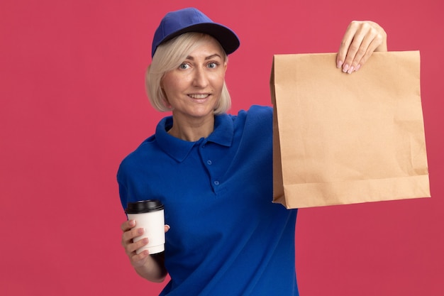 Smiling middle-aged blonde delivery woman in blue uniform and cap holding plastic coffee cup and paper package 