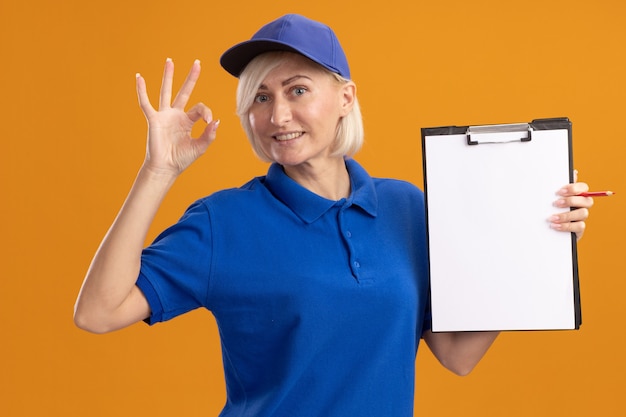 Улыбающаяся белокурая женщина-доставщик средних лет в синей униформе и кепке показывает буфер обмена на камеру, делает хорошо, знак изолирован на оранжевой стене