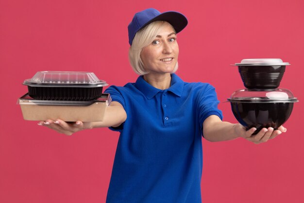 Smiling middle-aged blonde delivery woman in blue uniform and cap stretching out paper food package and food containers towards front looking at front isolated on pink wall