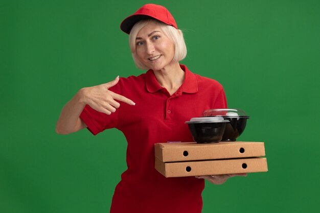 Sorridente donna di mezza età bionda consegna in uniforme rossa e berretto che tiene e indica i pacchetti di pizza con contenitori per alimenti su di essi isolati sul muro verde