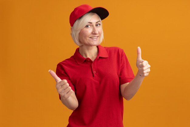 Улыбающаяся блондинка-курьерская женщина средних лет в красной форме и кепке показывает палец вверх