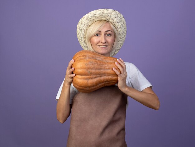 Улыбающаяся белокурая женщина-садовник средних лет в униформе в шляпе, держащая мускатную тыкву, смотрящую вперед, изолированную на фиолетовой стене с копией пространства