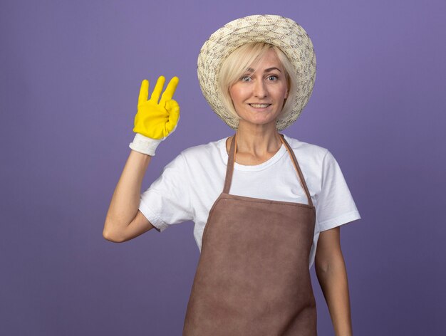 Smiling middle-aged blonde gardener woman in uniform wearing hat and gardening gloves doing ok sign 