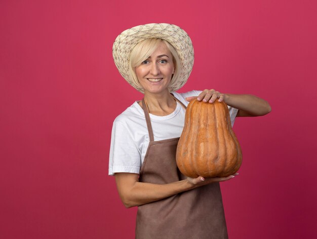 Улыбающаяся блондинка среднего возраста женщина-садовник в униформе в шляпе держит ореховую тыкву, изолированную на малиновой стене с копией пространства