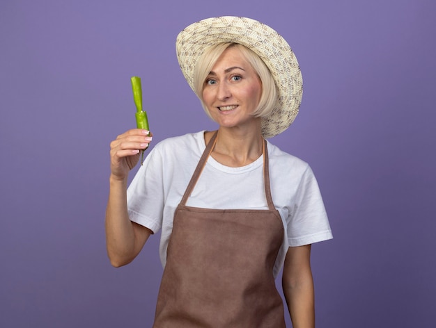 Smiling middle-aged blonde gardener woman in uniform wearing hat holding pepper looking at front isolated on purple wall with copy space