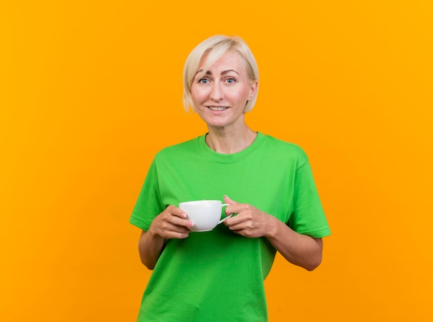 Smiling middle-aged blonde slavic woman holding cup of tea looking at front isolated on yellow wall with copy space