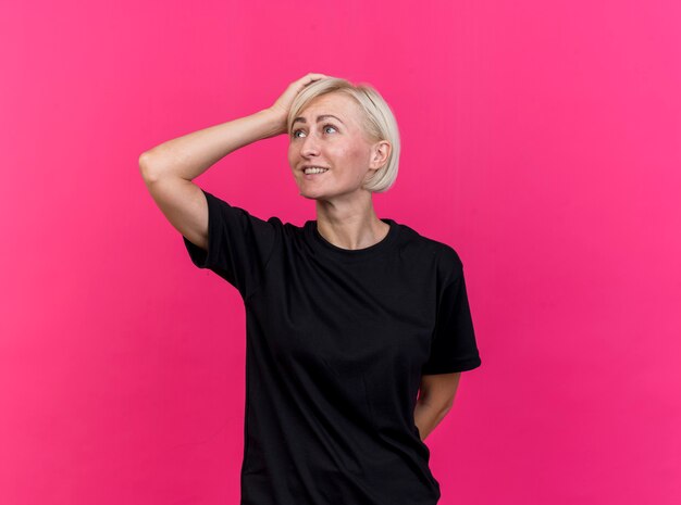 Smiling middle-aged blonde slavic woman keeping hands behind back and on head looking at side isolated on crimson background with copy space