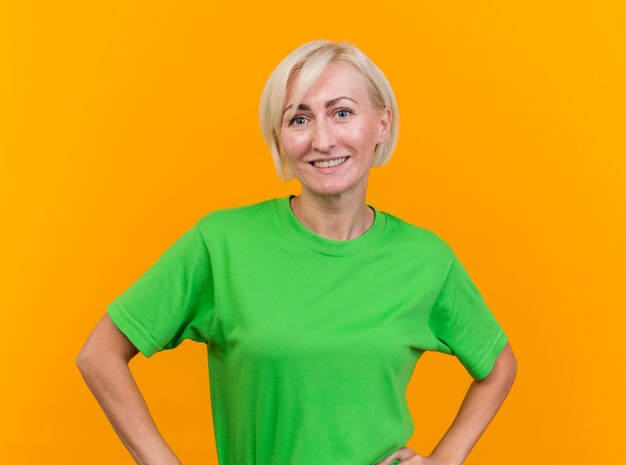 Smiling middle-aged blonde slavic woman looking at camera keeping hands on waist isolated on yellow background