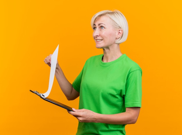 Smiling middle-aged blonde slavic woman standing in profile view holding clipboard looking straight isolated on yellow background