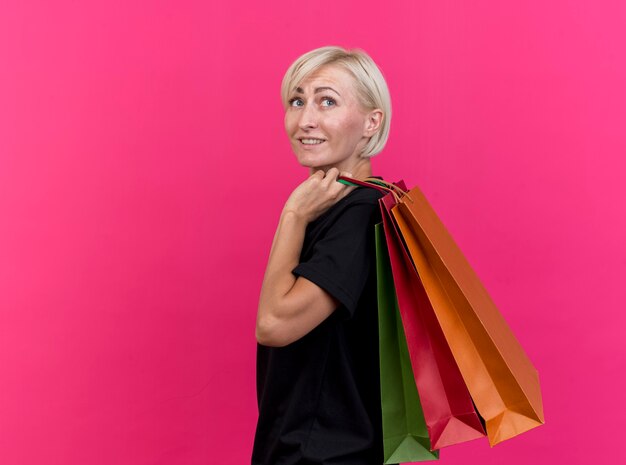 Smiling middle-aged blonde slavic woman standing in profile view holding shopping bags on shoulder looking at side isolated on crimson background with copy space
