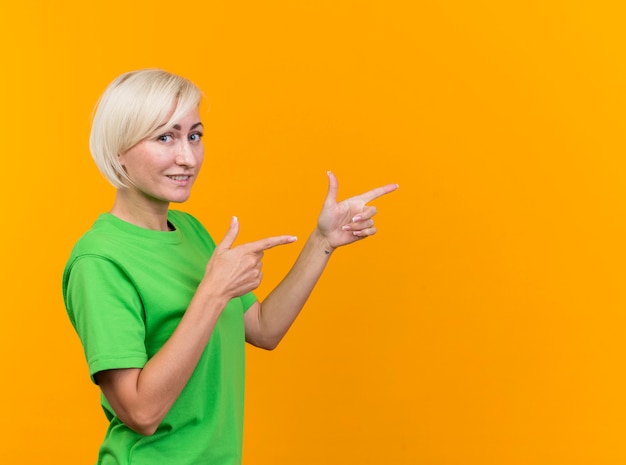 Smiling middle-aged blonde slavic woman standing in profile view looking at camera pointing straight isolated on yellow background with copy space