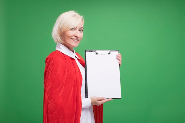 Smiling middle-aged blonde superhero woman in red cape standing in profile view looking at front showing clipboard to front isolated on green wall with copy space