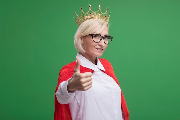 Free photo smiling middle-aged blonde superhero woman in red cape wearing glasses and crown  showing thumb up isolated on green wall with copy space