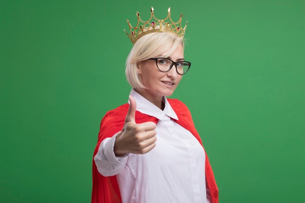 Smiling middle-aged blonde superhero woman in red cape wearing glasses and crown  showing thumb up isolated on green wall with copy space