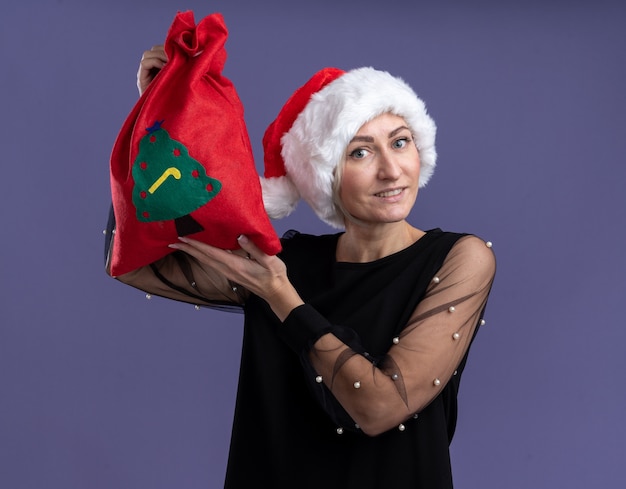 Smiling middle-aged blonde woman wearing christmas hat holding christmas sack near head looking at camera isolated on purple background