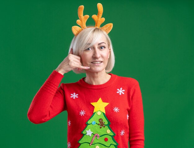 크리스마스 순록 뿔 머리띠와 크리스마스 스웨터를 입고 웃는 중년 금발의 여자가 복사 공간이 녹색 배경에 고립 된 전화 제스처를하고 카메라를보고