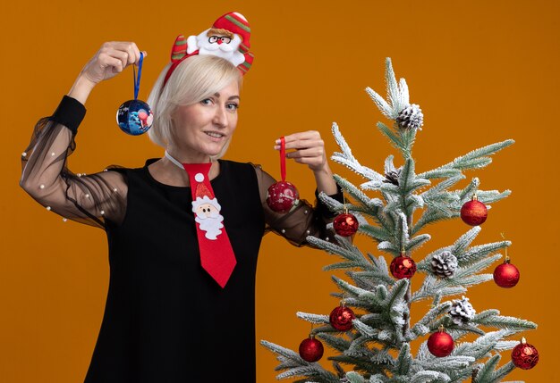 Smiling middle-aged blonde woman wearing santa claus headband and tie standing near decorated christmas tree holding christmas baubles looking at camera isolated on orange background