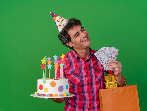 Smiling middle-aged caucasian party man wearing birthday cap holding birthday cake paper bag gift pack and money with closed eyes isolated on green background with copy space