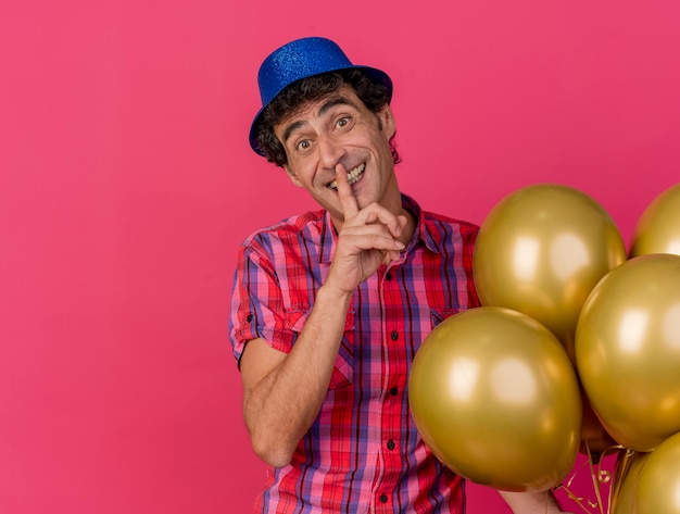 Smiling middle-aged caucasian party man wearing party hat holding balloons looking at camera doing silence gesture isolated on crimson background with copy space