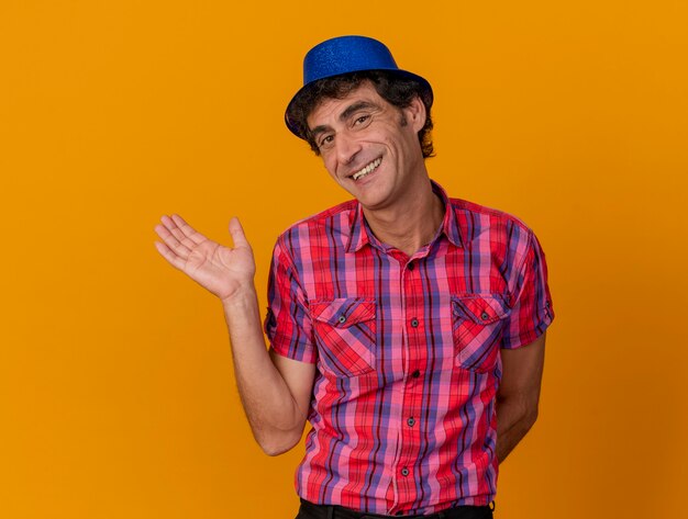 Smiling middle-aged caucasian party man wearing party hat looking at camera showing empty hand keeping another hand behind back isolated on orange background with copy space