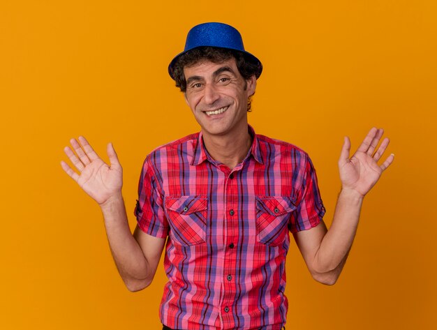 Smiling middle-aged caucasian party man wearing party hat looking at camera showing empty hands isolated on orange background
