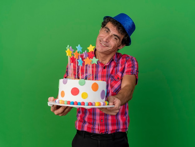 복사 공간이 녹색 배경에 고립 된 카메라를 향해 생일 케이크를 뻗어 카메라를보고 파티 모자를 쓰고 웃는 중년 백인 파티 남자