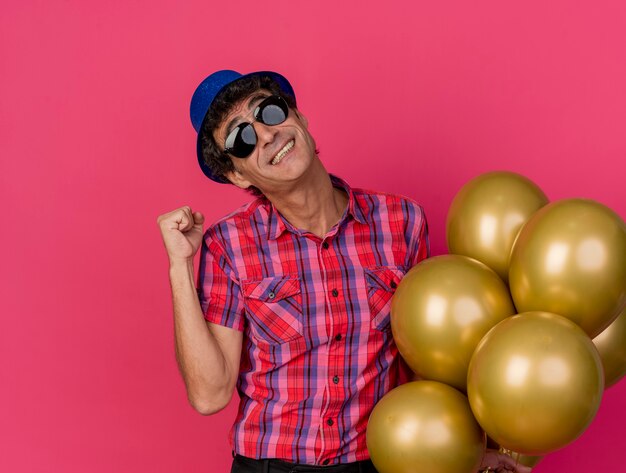 Smiling middle-aged caucasian party man wearing party hat and sunglasses holding balloons looking at camera doing yes gesture isolated on crimson background with copy space