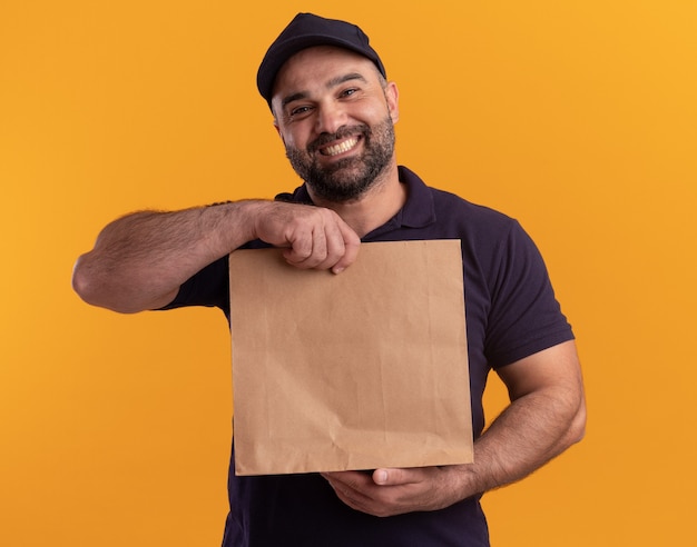 Улыбающийся курьер средних лет в униформе и кепке держит бумажный пакет с едой на желтой стене