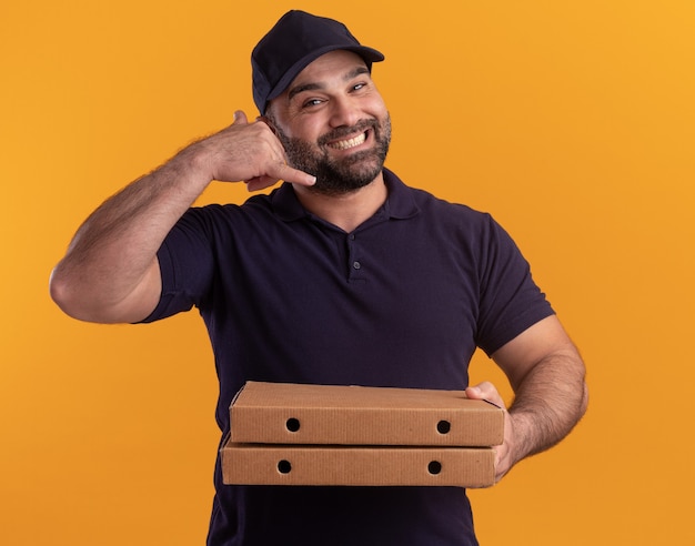 Smiling middle-aged delivery man in uniform and cap holding pizza boxes showing phone call gesture isolated on yellow wall