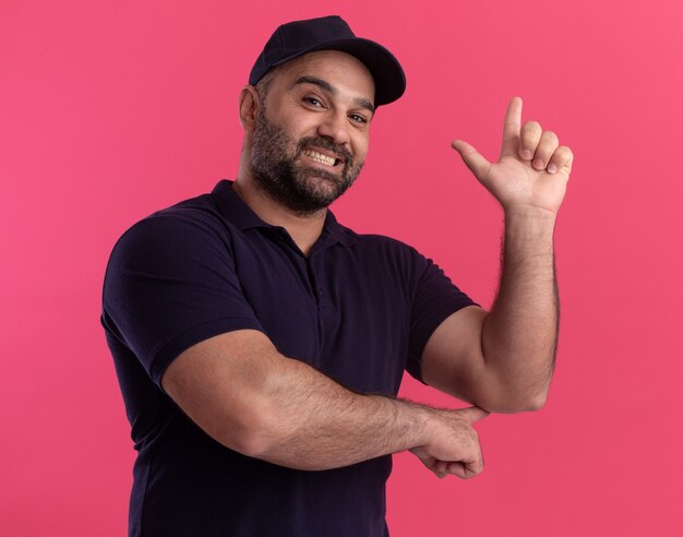 Smiling middle-aged delivery man in uniform and cap points at behind isolated on pink wall with copy spce