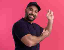 Free photo smiling middle-aged delivery man in uniform and cap points with hands at behind isolated on pink wall with copy space