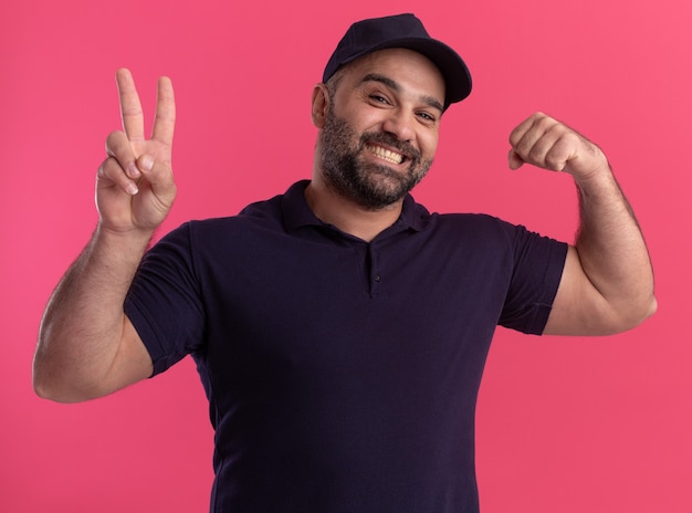 Smiling middle-aged delivery man in uniform and cap showing peace gesture and doing strong gesture isolated on pink wall