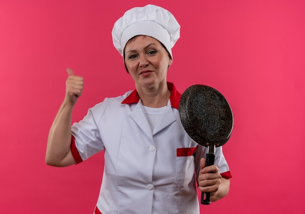 Smiling middle-aged female cook in chef uniform holding frying pan her thumb up