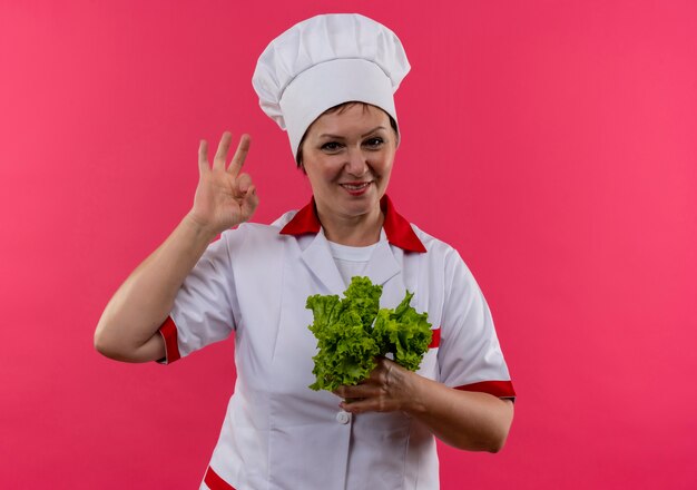 Smiling middle-aged female cook in chef uniform holding salad showing okey gesture