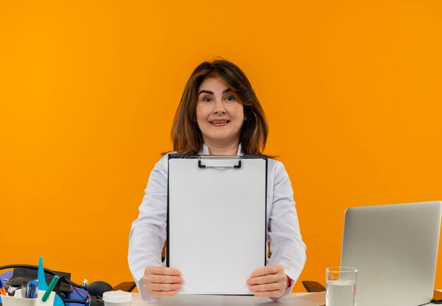 Smiling middle-aged female doctor wearing medical robe and stethoscope sitting at desk with medical tools and laptop showing clipboard isolated