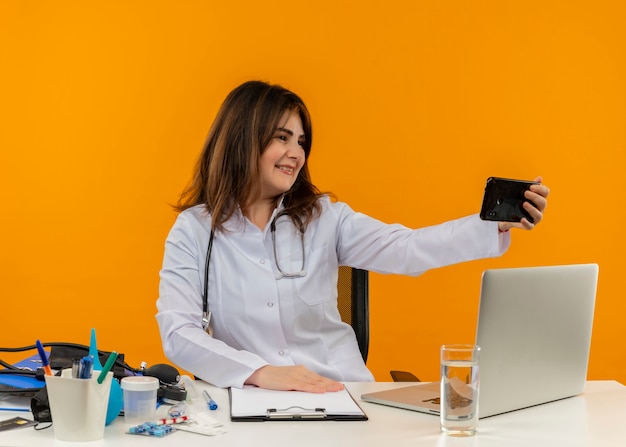 Улыбающаяся женщина-врач средних лет в медицинском халате со стетоскопом, сидя за столом, работает на ноутбуке с медицинскими инструментами, делает селфи на изолированном оранжевом фоне с копией пространства