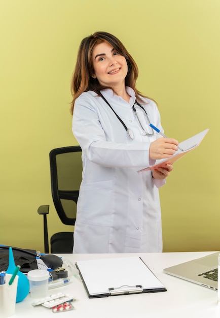 Улыбающаяся женщина-врач средних лет в медицинском халате со стетоскопом, сидя за столом, работает на ноутбуке с медицинскими инструментами, держа ручку и блокнот с копией пространства