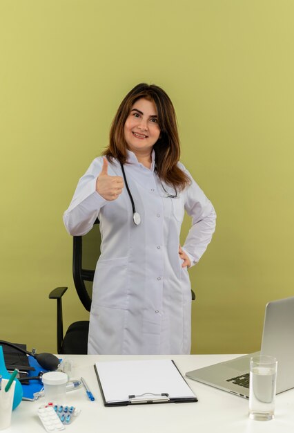 Smiling middle-aged female doctor wearing medical robe with stethoscope standing behind desk work on laptop with medical tools putting hand on hip her thumb up on green wall