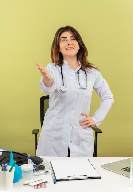 복사 공간이 카메라에 손을 들고 nedical 도구와 노트북에 책상 작업 뒤에 청진 서 의료 가운을 입고 웃는 중년 여성 의사