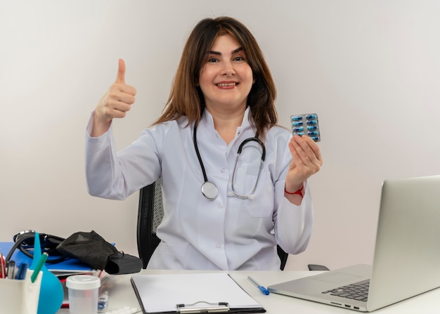 흰색 벽에 그녀의 엄지 손가락을 신용 카드를 들고 의료 도구와 노트북에 책상에 앉아 청진기와 의료 가운을 입고 입고 웃는 중년 여성 의사