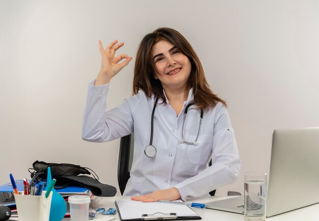 Smiling middle-aged female doctor wearing wearing medical robe with stethoscope sitting at desk work on laptop with medical tools showing okey gesture on isolated white backgroung with copy space