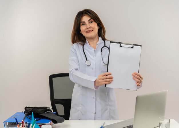 Smiling middle-aged female doctor wearing wearing medical robe with stethoscope standing behind desk work on laptop with medical tools holding clipboard on isolated white backgroung with copy space