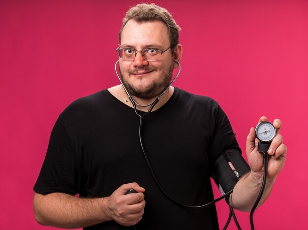 Smiling middle-aged ill male measuring his own pressure with sphygmomanometer isolated on pink wall