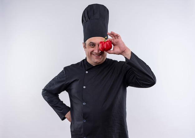 Free photo smiling middle-aged male cook in chef uniform covered eye with pepper putting his hand on hip