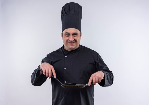 Smiling middle-aged male cook in chef uniform holding frying pan and spatula