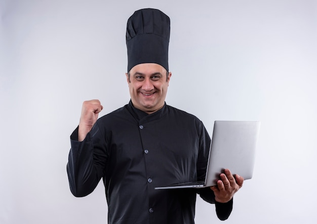 Smiling middle-aged male cook in chef uniform holding laptob showing yes gesture